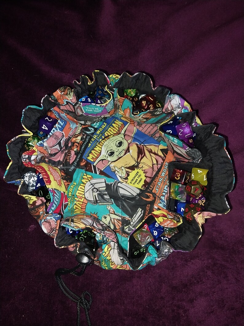 May include: A black fabric dice bag with a drawstring closure. The bag is decorated with a colourful comic book pattern featuring characters from Star Wars. The bag is filled with various coloured dice.