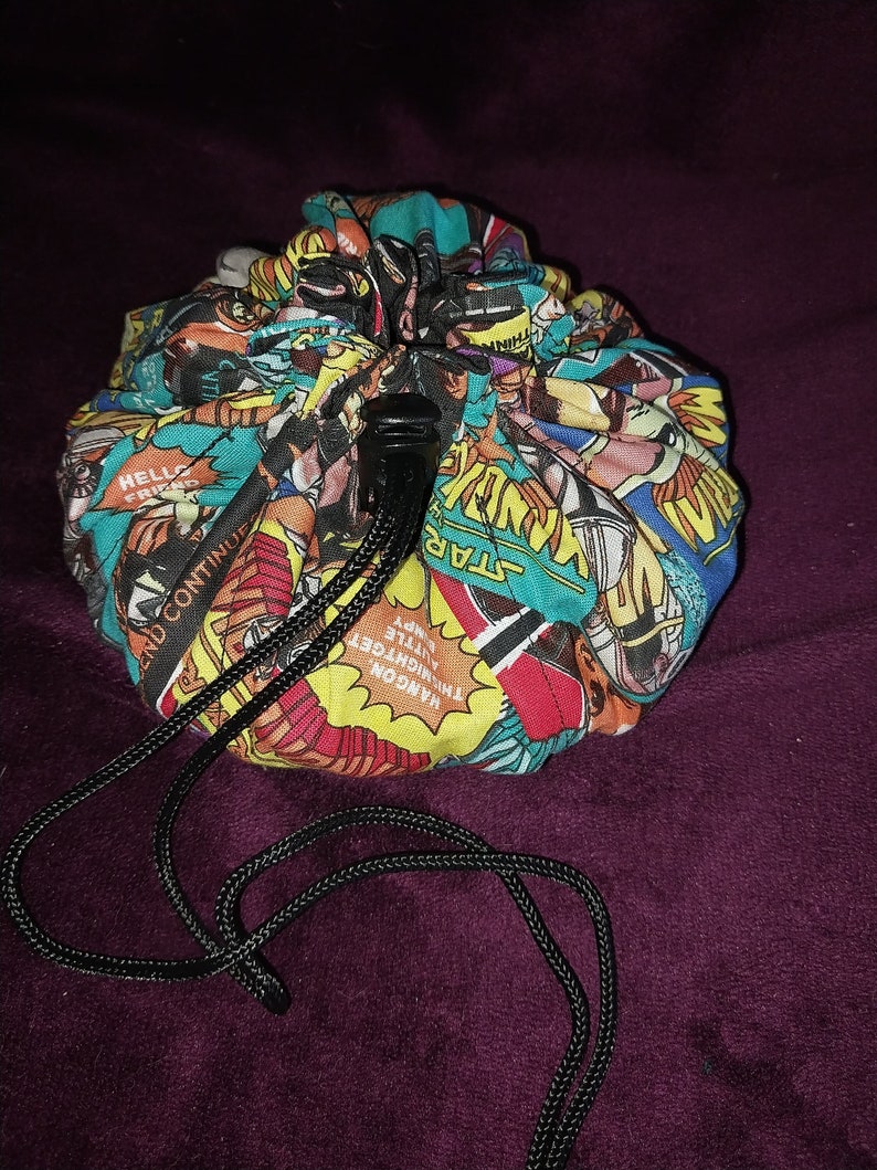 May include: A colourful fabric dice bag with a drawstring closure. The bag is decorated with a comic book pattern featuring superheroes and the words "Hangon", "The Midnight", "Little Early", "Star", and "And Continue".
