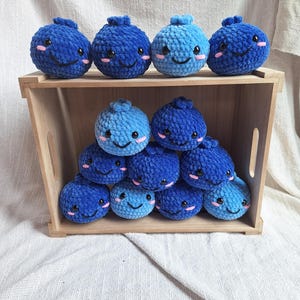 May include: A wooden crate filled with blue and light blue crocheted blueberry plush toys with smiling faces. The blueberries are arranged in a pyramid shape.
