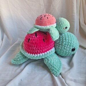 May include: Two crocheted plush turtle toys, one on top of the other. The larger turtle is green and the smaller turtle is pink and green, resembling a watermelon.