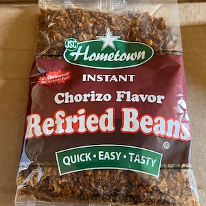 May include: A bag of instant chorizo flavor refried beans. The bag is green and white with the words "Hometown" and "Quick Easy Tasty" printed on it. The bag is filled with brown and red refried beans.
