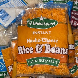 May include: Several packages of Hometown Instant Nacho Cheese Rice & Beans. The orange bags have the product name in large white letters. The bags also say "Quick, Easy, Tasty" and the net weight is 8 oz (227g). Other bags of rice are visible.