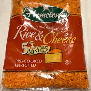 May include: A bag of orange-colored rice and cheese mix. The bag is labeled "Hometown Rice & Cheese" with a picture of a slice of cheese and a clock with the text "Ready in 5 Minutes". The bag also says "Long Grain" and "Pre-Cooked Enriched".