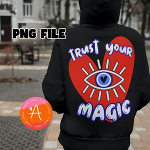 Può includere: Felpa nera con un design a forma di cuore rosso che presenta un occhio blu con pupilla nera e il testo "Trust Your Magic" in lettere bianche e blu.