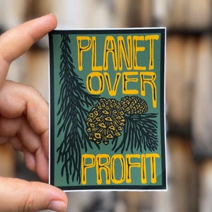 May include: A rectangular sticker with a green background and yellow text that reads "PLANET OVER PROFIT." The sticker features a pine branch and pine cones in black and yellow. The sticker is held by a hand.