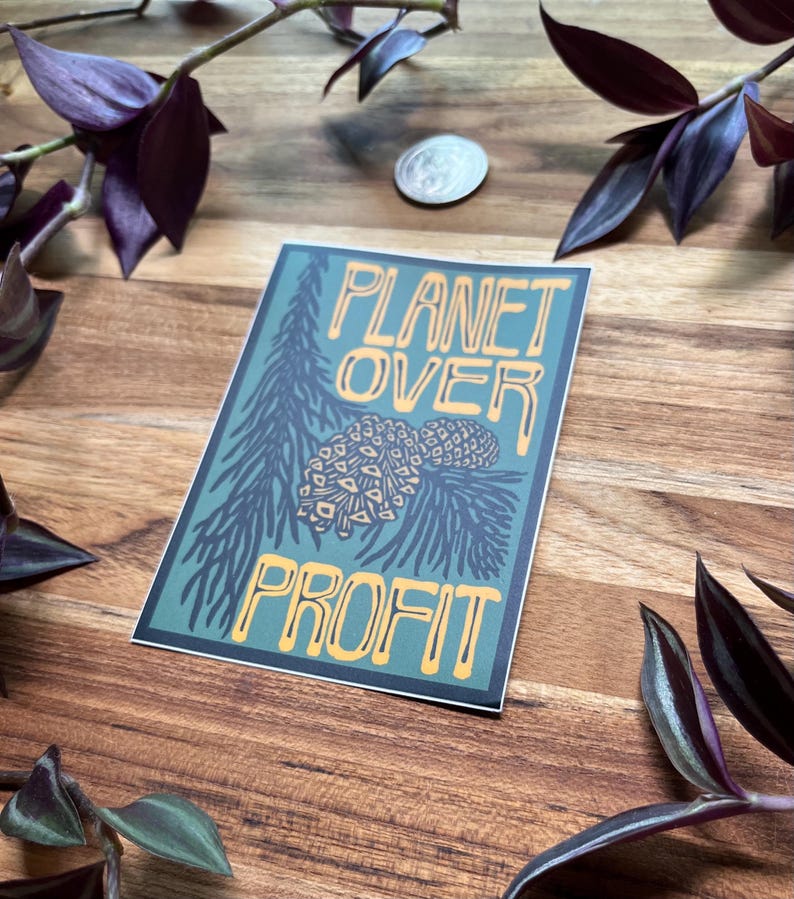 May include: A rectangular sticker with the words "PLANET OVER PROFIT" in yellow lettering. The sticker has a teal background with a pine tree and pine cone illustration. The sticker is on a wooden surface.