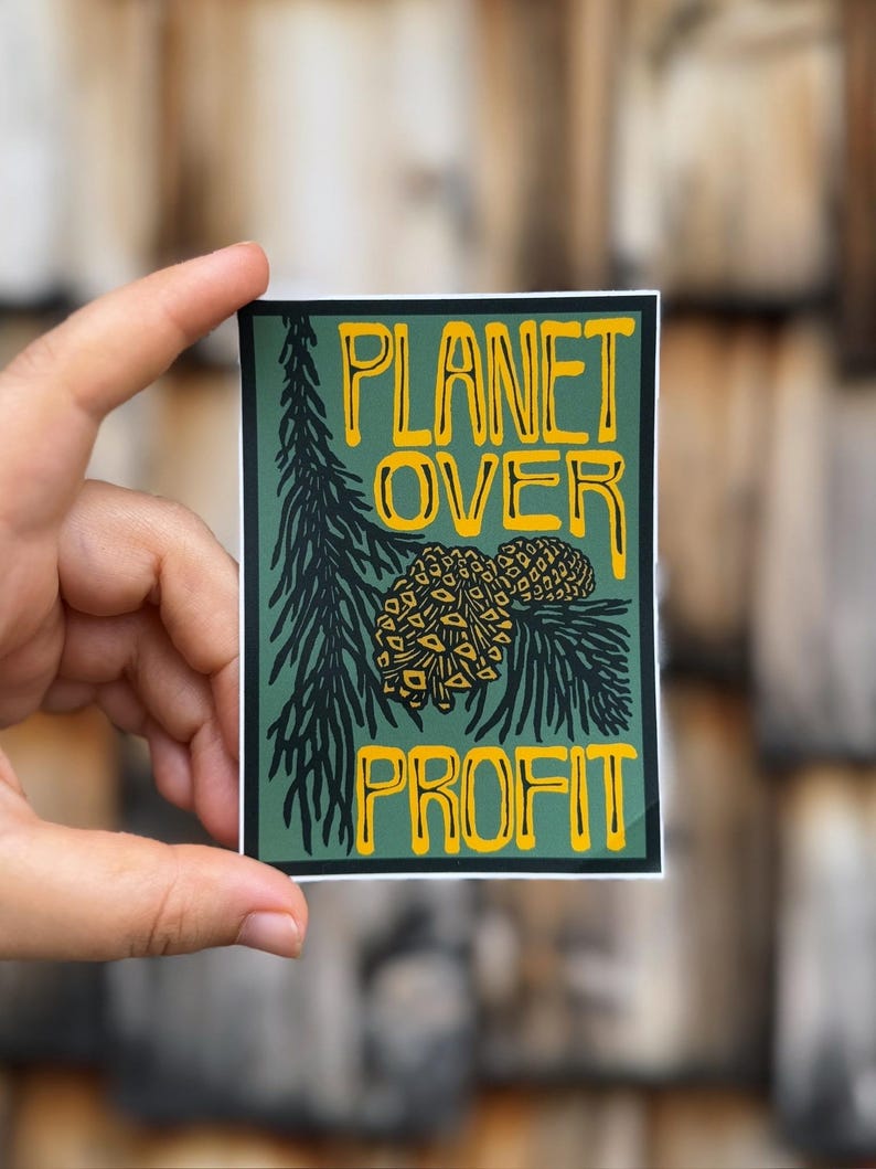 May include: A rectangular sticker with a green background and yellow text that reads "PLANET OVER PROFIT." The sticker features a pine branch and pine cones in black and yellow. The sticker is held by a hand.