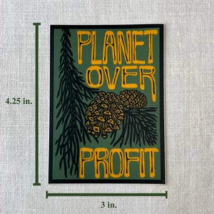 May include: A rectangular sticker with the text "PLANET OVER PROFIT" in yellow, set against a green background. The design includes a pine branch and two pinecones. The sticker measures 4.25 inches by 3 inches.