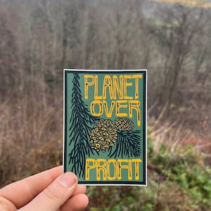 May include: A rectangular sticker with the text "PLANET OVER PROFIT" in yellow, outlined in black, on a dark green background. The design includes a pine branch and pine cones. The sticker is held up against a blurred outdoor background.