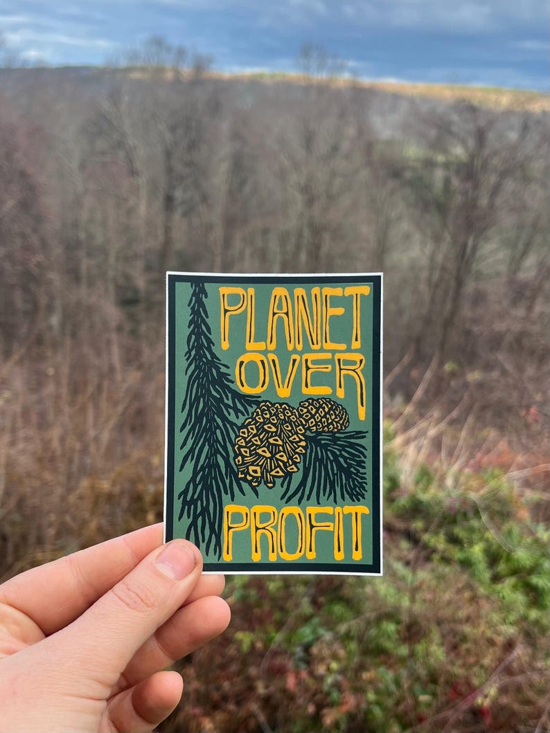 May include: A rectangular sticker with the text "PLANET OVER PROFIT" in yellow, outlined in black, on a dark green background. The design includes a pine branch and pine cones. The sticker is held up against a blurred outdoor background.