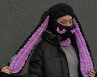 May include: A black knit hat with long purple ear flaps. The hat has a black knit face mask with a purple knit design.