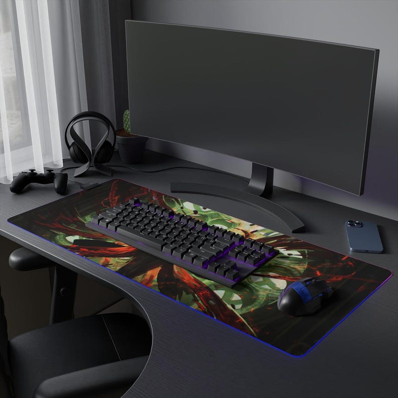 May include: A black and red gaming mouse pad with a colorful abstract design. A black keyboard with purple keys and a black gaming mouse are on the mouse pad. A black computer monitor and a black cell phone are on the desk.