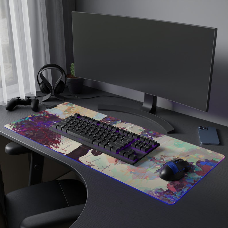 May include: A black mechanical keyboard, mouse, and smartphone rest on a large, colourful desk mat with an abstract design. A curved monitor, headphones, and a game controller are also visible. The mat has a blue LED edge.
