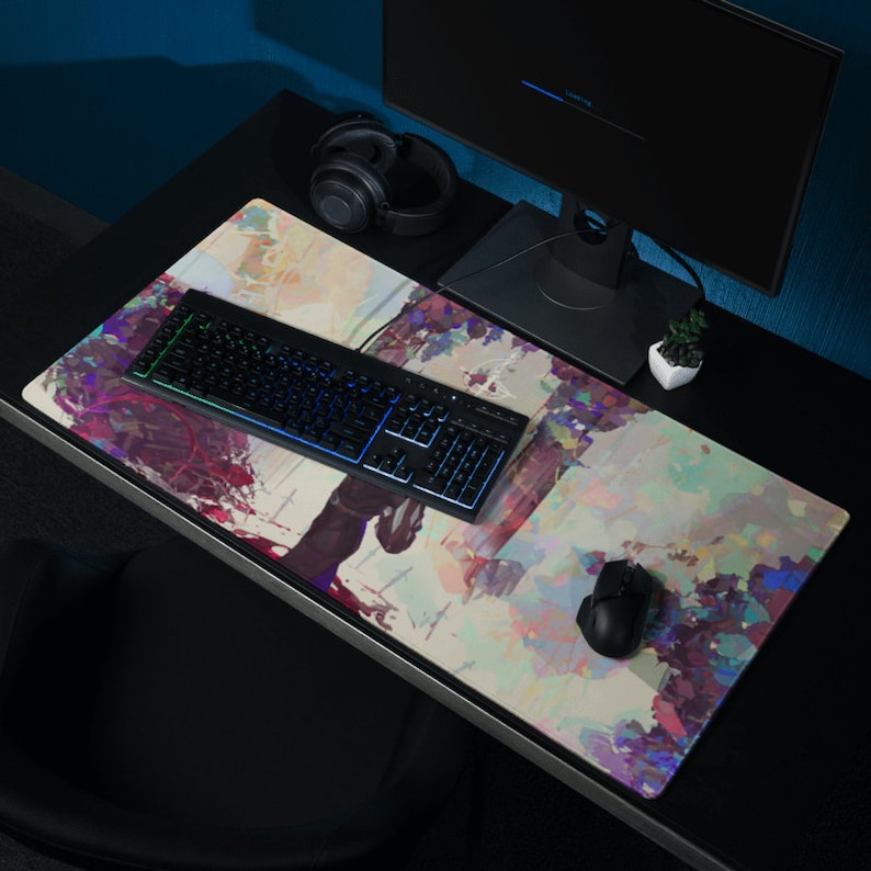 May include: A large rectangular mouse mat with an abstract watercolour design in shades of purple, pink, and green. A black keyboard with illuminated keys and a black mouse are on the mat. A computer monitor and headphones are also visible.