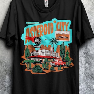 Puede incluir: Camiseta negra con un diseño gráfico retro que dice "WELCOME TO ASTEROID CITY". El diseño incluye un motel, un coche rojo, un paisaje desértico y un platillo volante. La camiseta está colgada en una percha negra.