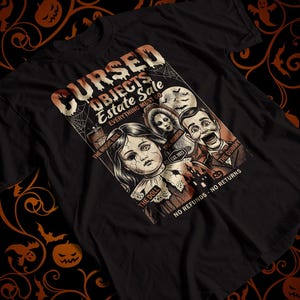 May include: A black short-sleeve t-shirt featuring a graphic print with the text CURSED OBJECTS ESTATE SALE EVERYTHING MUST GO. The design includes illustrations of a doll, a mirror, a music box, and a dummy, with the text THE DOLL, THE MIRROR, THE MUSIC BOX, THE DUMMY, and NO REFUNDS - NO RETURNS.