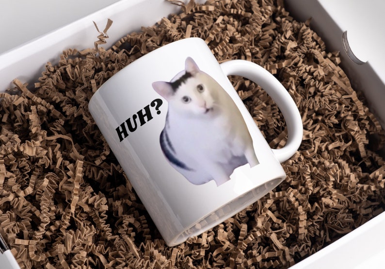 May include: White ceramic mug with a black and white cat image and the text "HUH?" printed on it. The mug is sitting in a brown paper shred box.
