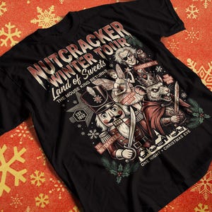 May include: A black short-sleeve t-shirt featuring a graphic print titled Nutcracker Winter Tour Land of Sweets. The design includes illustrations of a nutcracker, a mouse king with a sword, a fairy, and text including The Mouse King Returns.