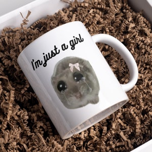 Può includere: Tazza in ceramica bianca con la scritta "I'm just a girl" e un'illustrazione di un criceto che piange con un fiocco rosa. La tazza è all'interno di una scatola bianca piena di carta crespa marrone.