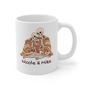 Può includere: Una tazza in ceramica bianca con un'illustrazione di due scheletri seduti insieme sotto una coperta a righe fantasia mentre tengono in mano una piccola tazza marrone. Sotto l'illustrazione, il testo Nicole & Mike è stampato in un font marrone scuro.