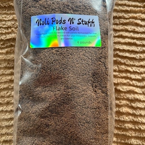 Peut inclure: Un sac en plastique transparent rempli de terreau en flocons brun. Le sac porte une étiquette qui indique "Noli Pods N' Stukk Flake Soil" et "Fabriqué à partir de granulés de chêne dur 100 % fermentés pendant plus de 45 jours. Nutrition complète pour vos mille-pattes, isopodes et scarabées. 6 livres."