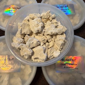 May include: A clear plastic container filled with beige limestone rubble and powder, surrounded by four similar containers. Each container has a label that reads "Noli Pods N' Stuff Limestone rubble & powder 8 oz."
