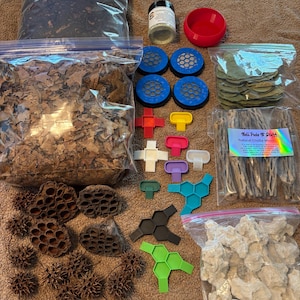 Op de afbeelding: Een assortiment terrariumbenodigdheden, waaronder zakken substraat, gedroogde bladeren en diverse decoratieve elementen. Er zijn ook kleine plastic componenten in blauw, groen, zwart en andere kleuren, evenals een rode kom.