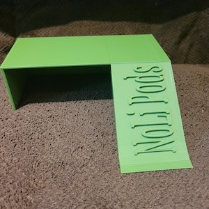 May include: A bright green, stepped platform with the text "Noli Pods" embossed on the side. The platform has a rectangular top and a lower, angled section, likely for display or storage.