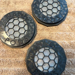 May include: Three dark gray, circular objects with the text "NOLI PODS" are displayed. Each has a geometric pattern around the edge and a honeycomb-like design in the center. The objects are on a textured, tan surface.