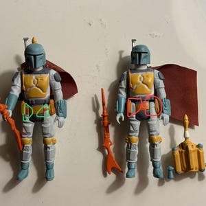 May include: Two Boba Fett action figures with blue-grey helmets, yellow chest armor, and grey jumpsuits. Each figure wears a dark red cape. One figure holds an orange blaster rifle, while the other stands next to a detached orange blaster and jetpack.