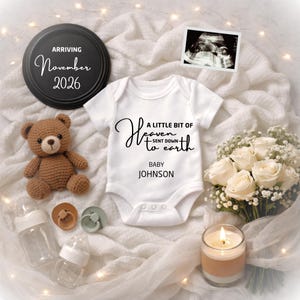Puede incluir: Anuncio de bebé con un body blanco con el texto "A LITTLE BIT OF Heaven sent down to earth BABY JOHNSON". La imagen incluye un osito de peluche, biberones, chupetes, una ecografía, rosas blancas y una vela.