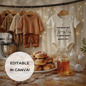 May include: An off-white baby onesie with the text "OUR BRAND NEW Honey Bun IS COMING SOON! JANUARY 2026" hangs on a wooden hanger. Several sets of baby clothes in neutral tones are on hangers. A plate of donuts, a jar of honey, and baby bottles are also visible.