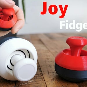以下が含まれることがあります： 画像には「Joy」と「Fidget」の文字が入った、赤、白、黒のフィジェットおもちゃ。おもちゃは円形のベースと中央の回転要素を持っています。上部は赤、ベースは黒、中央部分は白です。