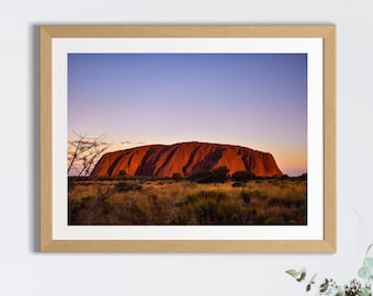 Op de afbeelding: Een ingelijste foto van Uluru, ook bekend als Ayers Rock, een grote zandstenen monoliet in het Northern Territory van Australië. De rots is roodbruin van kleur en wordt omringd door een grasveld. De lucht is lichtblauw met een vleugje roze in de verte.