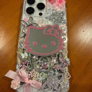 Op de afbeelding: Een doorzichtig telefoonhoesje versierd met diverse bedels, waaronder een groot Hello Kitty-gezicht, een zilveren planeetvormige bedel, een roze stoffen strik, een kleine roze roos, zilveren sterren, hartjes en vlinders met roze accenten.