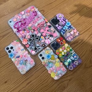 Könnte beinhalten: Eine Sammlung klarer Handy- und Tablet-Hüllen, die mit verschiedenen Verzierungen versehen sind. Die Hüllen weisen eine Vielzahl dekorativer Elemente auf, darunter Blumen, Herzen und das Wort "Believe". Die Hüllen sind auf einer Holzoberfläche angeordnet.