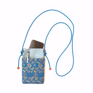 May include: A blue crossbody pouch featuring a gold floral vine pattern on the lower section and a solid light blue upper pocket. The bag includes a long blue rope strap with red knotted ends and contains a dark smartphone and a silver pen.