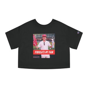 May include: Black cropped t-shirt with a photo of a person wearing a red baseball cap and holding a foam finger and a drink. The text "FIBULA'S #1 FAN" is printed in red below the photo.
