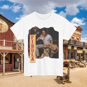 May include: White t-shirt with a graphic print of a man holding a beer and a dog. The graphic is set against a wooden background with the word "FIBULA" in red letters.