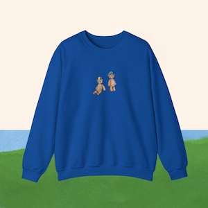 May include: A royal blue sweatshirt with a small embroidered design of two teddy bears wearing hats. One bear is wearing a yellow crown and the other is wearing a blue and green hat.