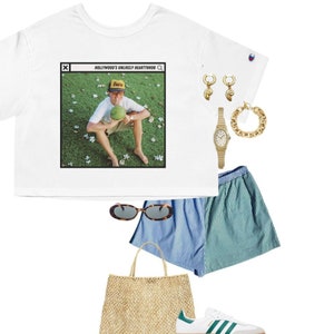 May include: A white cropped t-shirt with a graphic of a man wearing a yellow baseball cap and drinking from a coconut. The graphic is in a computer window with the text "HOLLYWOOD'S UNLIKELY HEARTTHROB" at the top. The shirt is paired with blue and green striped shorts, a straw tote bag, white sneakers with green stripes, tortoise shell sunglasses, a gold chain bracelet, and gold hoop earrings.