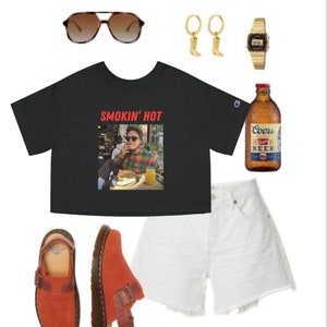 May include: A black cropped t-shirt with the text "Smokin' Hot" in red letters and a photo of a person wearing a plaid shirt and sunglasses. The t-shirt is paired with white denim shorts, red suede clogs, a pair of gold hoop earrings, a gold watch, and a bottle of Coors beer.