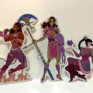 May include: Three fantasy-themed stickers featuring female characters in dynamic poses. The characters are depicted with glittery pink and purple outfits, and each holds a weapon or magical item. One sticker shows a character with an axe, another with a staff, and the third with a star-shaped object.