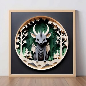 May include: A papercraft artwork depicting a gray, winged creature with a horn in a forest setting. The creature is surrounded by green trees and a crescent moon. The artwork is framed in a light wood frame.