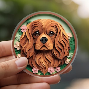 May include: A round sticker featuring a Cavalier King Charles Spaniel dog with brown fur and a black nose. The dog is surrounded by green leaves and pink flowers, set against a blurred background.