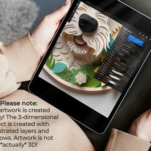 May include: A digital artwork of a dog's face is displayed on a tablet screen. The artwork is a 3D paper cut design with white, black, green, and blue elements. The tablet is held by a person wearing a tan sweater. The text on the screen says, "Please note: My artwork is created digitally! The 3-dimensional effect is created with illustrated layers and shadows. Artwork is not *actually* 3D!"