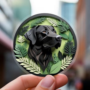 May include: A round sticker featuring a black Labrador Retriever dog portrait surrounded by green leaves. The dog's head is in profile, with detailed fur and brown eyes. The sticker has a transparent border.