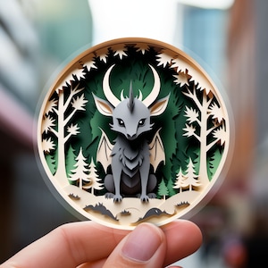 May include: A papercraft sculpture of a grey, winged creature with horns and a pointed snout, set against a green forest background. The sculpture is detailed with intricate paper cuts and is displayed in a circular frame.