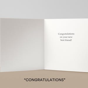 May include: An open greeting card with the text "Congratulations on your new best friend!" on the right panel. The bottom of the image displays the word "CONGRATULATIONS" in quotation marks. The card is white with a neutral background.