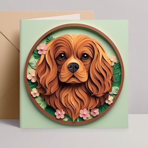 May include: A square greeting card featuring a detailed paper-cut illustration of a Cavalier King Charles Spaniel dog within a circular frame. The dog is brown with long, wavy fur, surrounded by pink flowers and green leaves. A brown envelope is to the left.